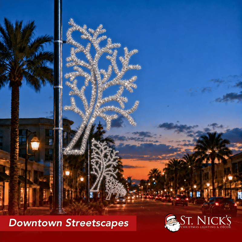 A downtown boulevard at sunset is lined with silver Winter Branch Tree Tinsel decorations mounted on black light poles. The repeating tree-shaped displays create a coordinated winter streetscape along the palm-lined commercial road.