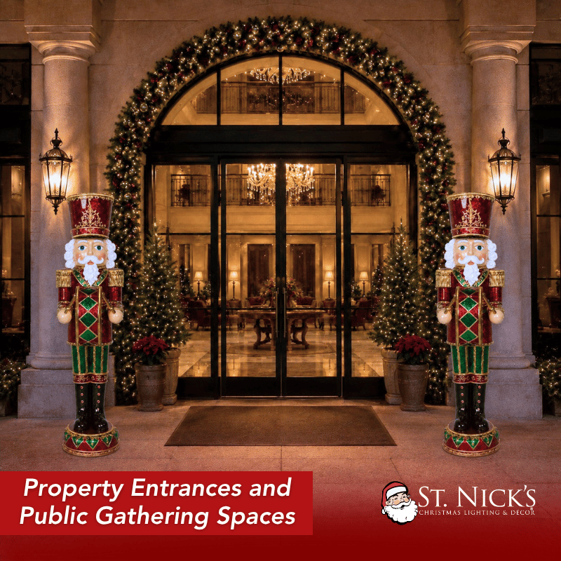 A matching pair of 6-foot Toy Soldier Nutcrackers displayed on either side of an elegant property entrance decorated with a large illuminated garland arch, wreaths, poinsettias, Christmas trees, and warm evening lighting.