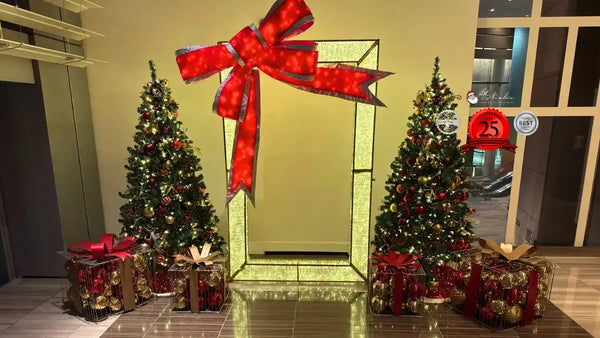 Decorative Christmas scene with two trees, presents, and a large red bow in front of a door.
