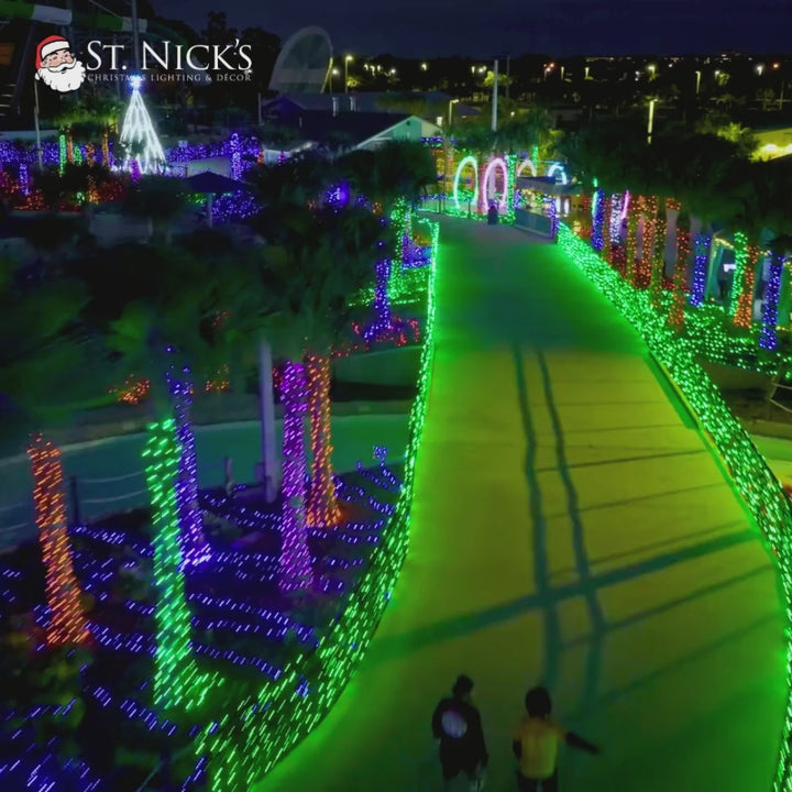 Aerial video tours a colorful holiday display with illuminated palms, landscaped beds, arches, and glowing pathways.