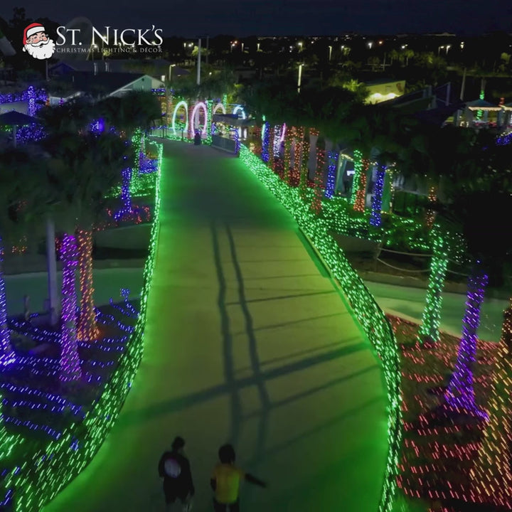 Nighttime aerial view of a large commercial holiday attraction illuminated with green, purple, blue, and other vibrant lights.