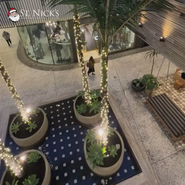 Video looks over indoor palm trees wrapped in warm white LED mini lights throughout a modern commercial plaza.